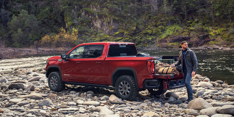 A man stands at the open tailgate of his red 2026 GMC Sierra 1500 which is parked on the rocky shore of a river beside a forested cliff
